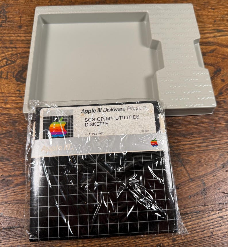 May include: A vintage Apple III Diskware Program SOS-CP/M Utilities diskette in its original plastic wrap, featuring a black grid pattern and the classic rainbow Apple logo. It is accompanied by a light grey plastic storage tray with Apple branding.