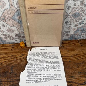 May include: A tan software manual titled Catalyst for the Apple III by Quark, enclosed in a clear plastic binder with a three-hole punched spine. A separate white paper sheet with black text titled DEALERS is included, detailing software usage.