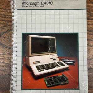 K&ouml;nnte beinhalten: Ein spiralgebundenes Apple III Microsoft BASIC Referenzhandbuch mit wei&szlig;em Einband und Gittermuster. Das Cover zeigt ein Bild eines Apple III Computers, drei Disketten und eine Leiterplatte auf einer dunkelroten Oberfl&auml;che.
