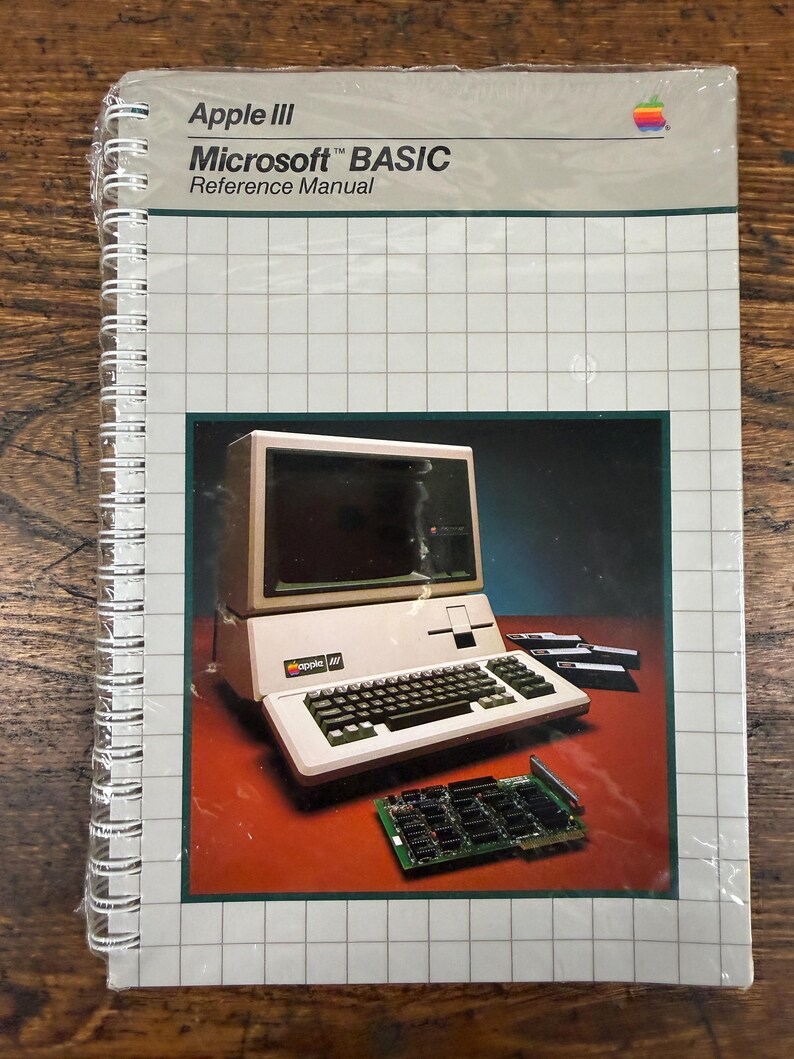 K&ouml;nnte beinhalten: Ein spiralgebundenes Apple III Microsoft BASIC Referenzhandbuch mit wei&szlig;em Einband und Gittermuster. Das Cover zeigt ein Bild eines Apple III Computers, drei Disketten und eine Leiterplatte auf einer dunkelroten Oberfl&auml;che.