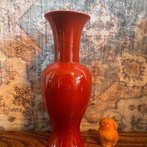 May include: A tall, glossy, solid red ceramic vase with a flared rim and a rounded body, positioned next to a small, carved wooden figurine of a dog with a light brown finish and simple, hand-painted facial features.