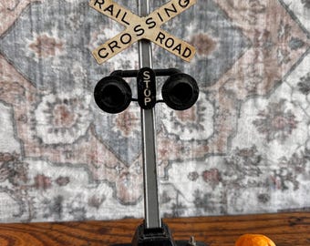 May include: A miniature metal railroad crossing signal with a black base, a vertical post, and a crossbuck sign reading RAIL CROSSING ROAD. Below the sign are two black circular lights and a small vertical sign reading STOP. Beside it sits a small orange dog figurine.