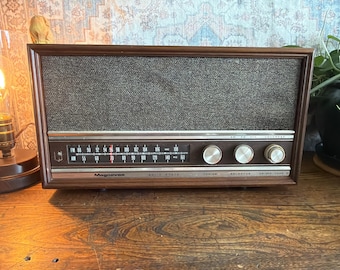 May include: Vintage Magnavox solid state radio with a dark wood frame and a gray speaker cover. The radio features a tuning dial, selector knobs, and a frequency display. The radio is sitting on a wooden surface.