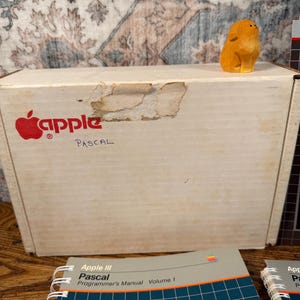 Puede incluir: Una caja de cart&oacute;n blanca con el logotipo rojo de Apple y la palabra PASCAL escrita en tinta negra. Una peque&ntilde;a figura de animal de madera tallada de color naranja se encuentra sobre la caja. En primer plano, dos manuales Apple III Pascal en espiral.