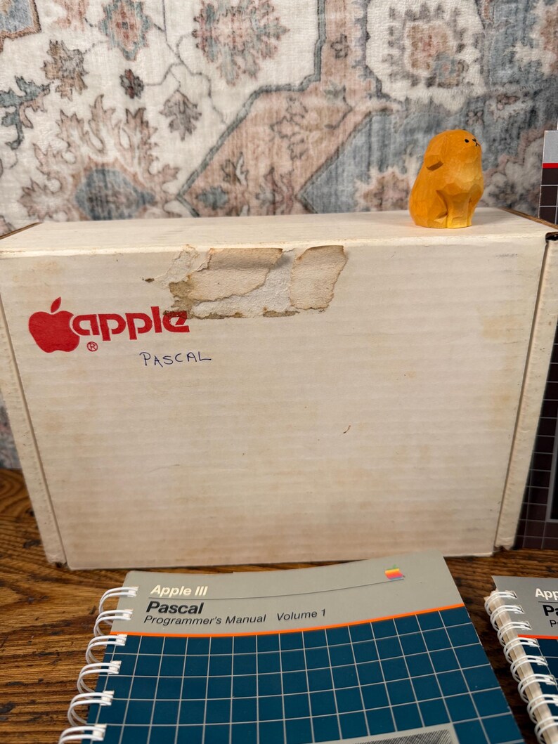 Puede incluir: Una caja de cart&oacute;n blanca con el logotipo rojo de Apple y la palabra PASCAL escrita en tinta negra. Una peque&ntilde;a figura de animal de madera tallada de color naranja se encuentra sobre la caja. En primer plano, dos manuales Apple III Pascal en espiral.