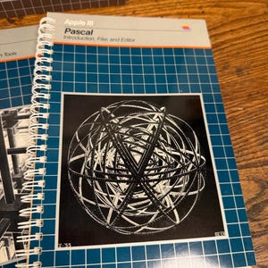 Puede incluir: Un manual de Apple III Pascal con encuadernaci&oacute;n en espiral y cubierta de cuadr&iacute;cula azul que presenta un gr&aacute;fico en blanco y negro de una esfera geom&eacute;trica compleja. El texto dice Apple III Pascal Introduction, Filer, and Editor con el logo.