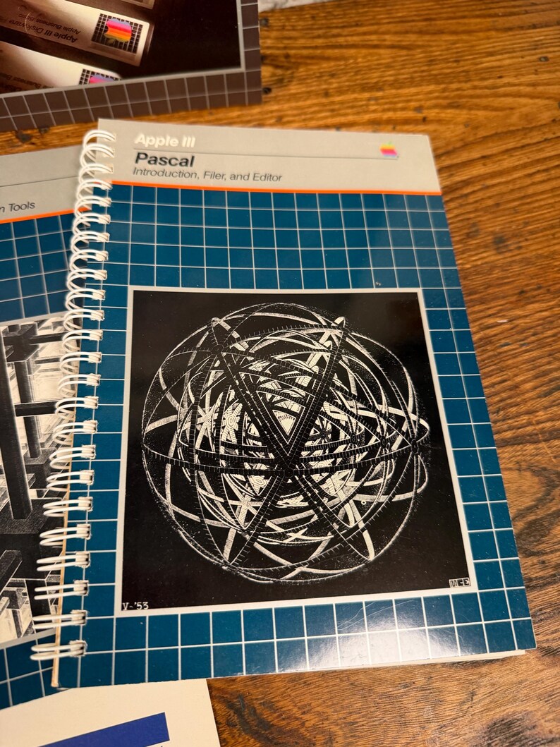 Puede incluir: Un manual de Apple III Pascal con encuadernaci&oacute;n en espiral y cubierta de cuadr&iacute;cula azul que presenta un gr&aacute;fico en blanco y negro de una esfera geom&eacute;trica compleja. El texto dice Apple III Pascal Introduction, Filer, and Editor con el logo.
