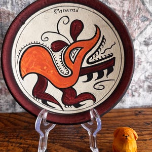 May include: A round ceramic plate with a dark red rim and a cream-colored center featuring a stylized orange and dark red creature illustration and the text Panama. A small, carved orange wooden dog figurine sits to the right of the plate.