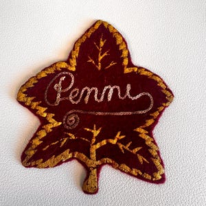 May include: A dark red felt leaf-shaped patch featuring gold embroidered edges and a gold embroidered tree design at the bottom. The name "Penne" is stitched in light pink thread across the center of the leaf in cursive script.