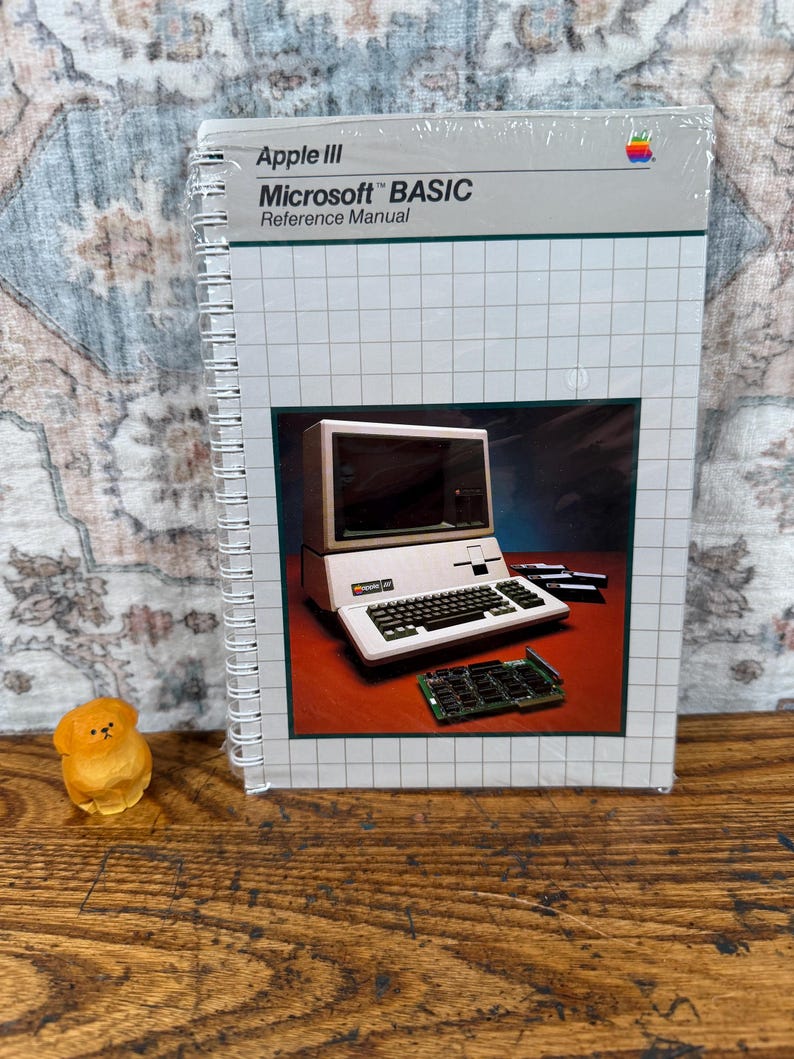 K&ouml;nnte beinhalten: Ein spiralgebundenes Apple III Microsoft BASIC Referenzhandbuch mit wei&szlig;em Einband, Gittermuster und einem zentralen Foto eines Apple III Computers, einer Tastatur und einer Leiterplatte. Das Apple-Logo befindet sich oben rechts.