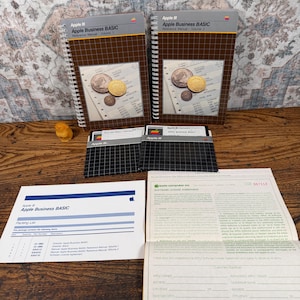 May include: Two spiral-bound Apple III Apple Business BASIC reference manuals, two black grid-patterned floppy disks, a white packing list document, and a beige software license agreement form. The manuals feature a brown grid cover design.
