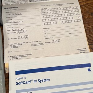 May include: A white Apple III SoftCard III System packing list document with blue horizontal lines and text, positioned below a beige Apple Computer limited warranty form with black text and fields for product information and customer details.