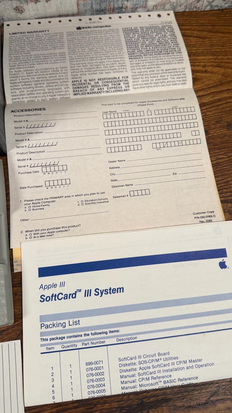 May include: A white Apple III SoftCard III System packing list document with blue horizontal lines and text, positioned below a beige Apple Computer limited warranty form with black text and fields for product information and customer details.