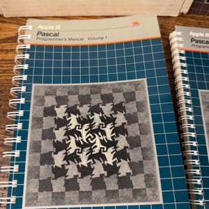 Puede incluir: Un manual de programaci&oacute;n Apple III Pascal Volumen 1 con encuadernaci&oacute;n en espiral, cubierta azul oscuro con patr&oacute;n de cuadr&iacute;cula blanca y un gr&aacute;fico cuadrado central de figuras de lagartos entrelazadas en blanco y negro. Logo Apple visible.