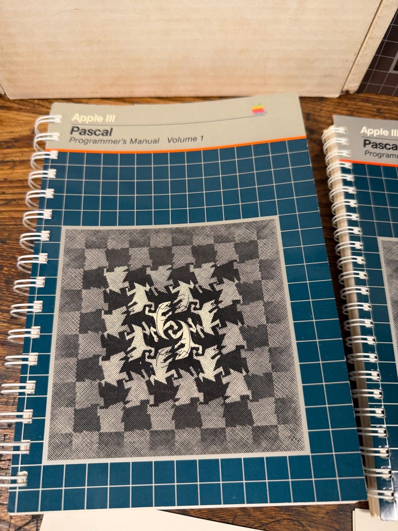 Puede incluir: Un manual de programaci&oacute;n Apple III Pascal Volumen 1 con encuadernaci&oacute;n en espiral, cubierta azul oscuro con patr&oacute;n de cuadr&iacute;cula blanca y un gr&aacute;fico cuadrado central de figuras de lagartos entrelazadas en blanco y negro. Logo Apple visible.