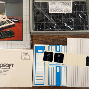 May include: A vintage Microsoft software package including a manual with a computer illustration, a plastic-wrapped diskette box, a white envelope with Microsoft Consumer Products branding, and a sheet of black drive letter stickers labelled A, B, E, and F.