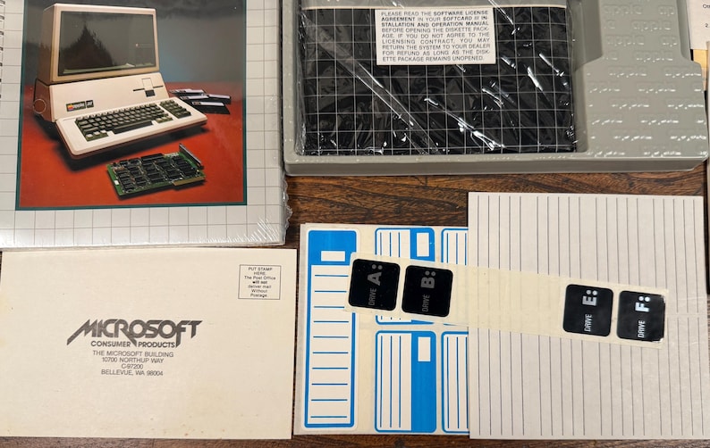 May include: A vintage Microsoft software package including a manual with a computer illustration, a plastic-wrapped diskette box, a white envelope with Microsoft Consumer Products branding, and a sheet of black drive letter stickers labelled A, B, E, and F.