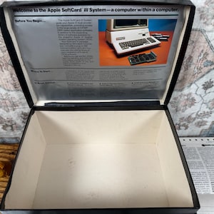 May include: An open black storage box with a white interior. The inside lid features a printed manual titled Welcome to the Apple SoftCard III System&mdash;a computer within a computer, including text and a photograph of an Apple computer and circuit board.