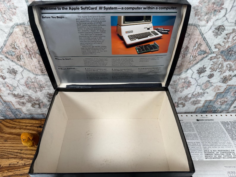 May include: An open black storage box with a white interior. The inside lid features a printed manual titled Welcome to the Apple SoftCard III System&mdash;a computer within a computer, including text and a photograph of an Apple computer and circuit board.
