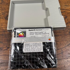 May include: A vintage Apple III Diskware Program master diskette in a clear plastic sleeve, featuring a rainbow Apple logo and grid pattern. The label reads Apple Softcard III CP/M Master Diskette, accompanied by a light grey plastic storage tray.