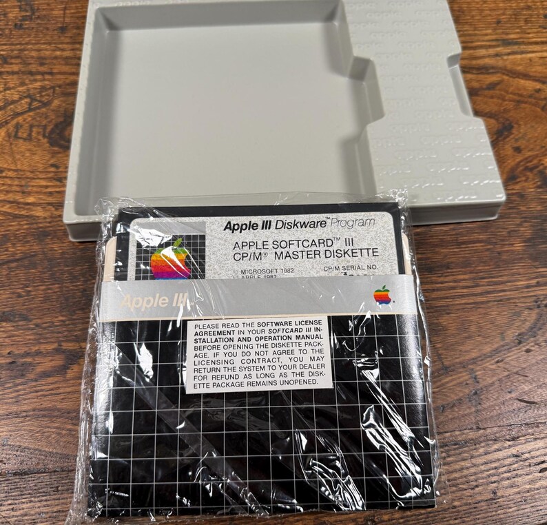 May include: A vintage Apple III Diskware Program master diskette in a clear plastic sleeve, featuring a rainbow Apple logo and grid pattern. The label reads Apple Softcard III CP/M Master Diskette, accompanied by a light grey plastic storage tray.