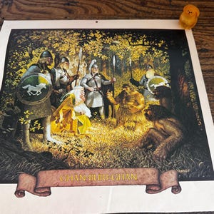 May include: A rectangular print featuring a fantasy illustration of armored warriors and wild figures in a golden forest. A banner at the bottom displays the text GHAN-BURI-GHAN. A small yellow figurine sits in the top right corner of the print.