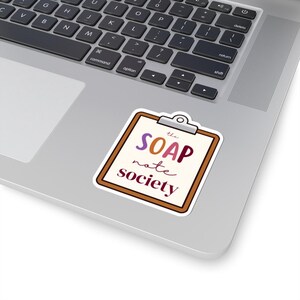 May include: A die-cut sticker shaped like a brown clipboard with a silver clip at the top. The center features the text 'the SOAP note society' in a mix of purple, orange, and dark red lettering against a cream-colored background.
