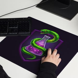 May include: A dark purple rectangular mousepad with a green snake coiled around a purple potion bottle design. The mousepad is on a white desk next to a black keyboard. The design is set against a purple shield.