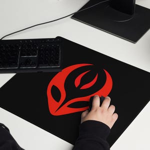 May include: Black rectangular mousepad with a red abstract design. The mousepad is on a white desk next to a black keyboard and a computer monitor. A person is using the mouse.