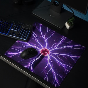 Op de afbeelding: Een zwarte computermuis en toetsenbord liggen op een zwart bureau, naast een paarse muismat met een plasma bal ontwerp. Een computermonitor en koptelefoon zijn ook aanwezig. De muismat heeft een centrale rode bol met uitstralende paarse bliksemschichten.