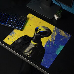 Puede incluir: Alfombrilla de ratón con diseño de alienígena con ojos azules sobre fondo amarillo y azul. La alfombrilla está sobre un escritorio negro con teclado, monitor y auriculares. El diseño tiene salpicaduras de pintura azul.