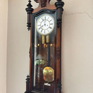 Puede incluir: Un reloj de abuelo antiguo de madera oscura con detalles negros. El reloj tiene una esfera blanca con números romanos, una puerta de cristal y un péndulo. La altura del reloj es de aproximadamente 180 cm y está contra una pared beige.