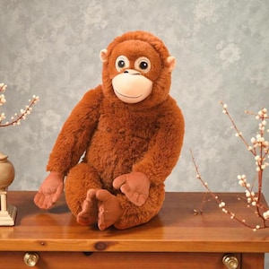 May include: A soft toy orangutan with brown fur, big eyes, and a pale face. The plush animal is seated on a wooden surface. The toy has a friendly expression and is designed to resemble a real orangutan.