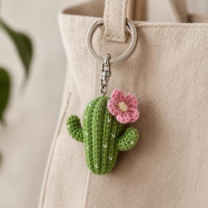 May include: A green crochet cactus keychain with a pink flower and white accents. The cactus has two arms and is attached to a silver metal keyring. The keychain is hanging on a beige tote bag.