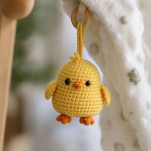 Può includere: Un giocattolo a forma di pulcino giallo fatto a mano all'uncinetto, con zampe e becco arancioni e occhi neri. Il pulcino è sospeso a un anello di legno tramite un anello all'uncinetto giallo. Lo sfondo include una coperta bianca.