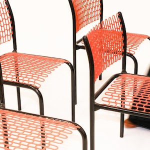 May include: A set of four chairs featuring black metal frames and bright orange seats and backrests constructed from a repeating, interconnected wire mesh pattern. The chairs have a simple, four-legged design with a curved back support structure.