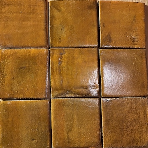 May include: A grid of nine square tiles arranged in three rows and three columns. Each tile features a glossy, mustard-yellow glaze with subtle variations in tone, visible surface texture, and slight imperfections that create a rustic appearance.