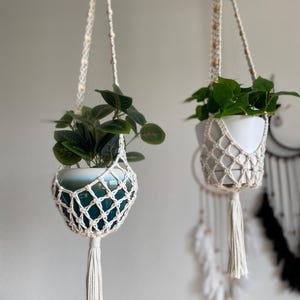 May include: Two cream-coloured macrame plant hangers, each holding a white pot with a green plant. The hangers are made of woven rope, with a net design to cradle the pots. Long tassels hang from the bottom, giving a bohemian feel.