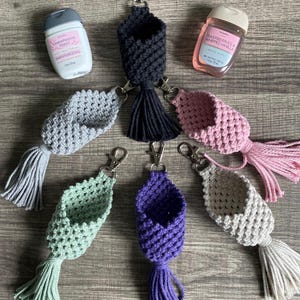 May include: Six macrame hand sanitizer holders in black, gray, pink, light green, purple, and beige, each with a metal lobster clasp and tassel. Two hand sanitizer bottles are included, one labeled Champagne Toast and one Raspberries & Whipped Vanilla.