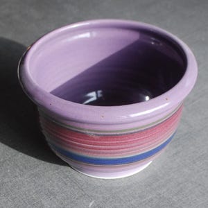 Può includere: Una ciotola in ceramica con una finitura liscia e lucida. L'interno e il bordo superiore sono di un colore viola chiaro uniforme. L'esterno presenta bande orizzontali di colore che includono tonalità di rosa, blu e verde spento, con base bianca.