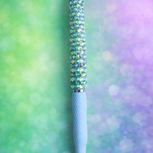 May include: A decorative pen with a light blue, textured grip and a barrel covered in iridescent rhinestones. The pen has a silver tip and is set against a blurred green and purple background.