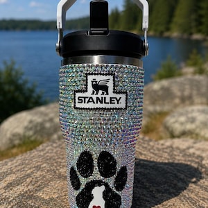 Op de afbeelding: Een witte Stanley-beker bedekt met iriserende strass-steentjes met een zwart Stanley-logo en een groot zwart pootafdrukontwerp. De pootafdruk bevat een wit silhouet van een hond en een klein rood hartje. Het heeft een zwarte deksel en een witte handgreep.