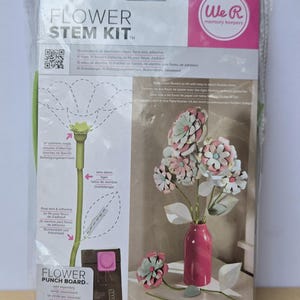 Könnte beinhalten: Ein Blumenstiel-Kit in einer transparenten Kunststoffverpackung. Die Verpackung trägt die Aufschrift "FLOWER STEM KIT" und "We R Memory Keepers". Das Bild zeigt eine rosa Vase mit Papierblumen und grünen Stielen.