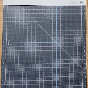 May include: A dark gray Cricut cutting mat with a grid pattern and measurements in inches. The mat has a white top with product information and a hole for hanging. The grid is marked with numbers from 0 to 24.
