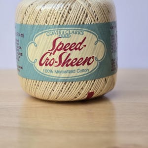 May include: A spool of beige Speed-Cro-Sheen thread by Coats & Clark's. The label is teal with the brand name in red script. The thread is 100% mercerized cotton. The spool is on a light wood surface.