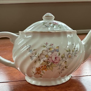 Puede incluir: Una tetera de porcelana blanca con textura acanalada en espiral y tapa a juego. El cuerpo presenta un delicado patrón floral con rosas rosadas, pequeñas flores azules y naranjas, y hojas verdes, acentuado con un fino borde dorado en los bordes.