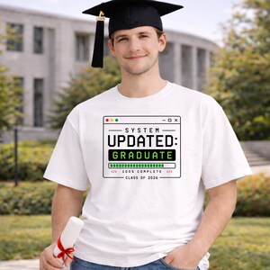 May include: White t-shirt with the text "SYSTEM UPDATED: GRADUATE" in a computer window design. The shirt also includes "100% COMPLETE" and "CLASS OF 2026". A person wearing a graduation cap and holding a diploma.