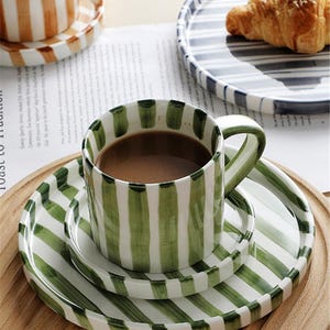 May include: A green and white striped ceramic coffee cup and saucer set with a matching plate. The cup is filled with coffee. A second cup and saucer set with brown and white stripes is visible in the background. A plate with a croissant is also visible.