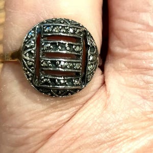 Puede incluir: Anillo vintage con diseño de filigrana plateada. El anillo presenta una piedra rectangular de color rojo anaranjado enmarcada por un engaste calado y detallado. La banda es dorada. Una joya clásica.