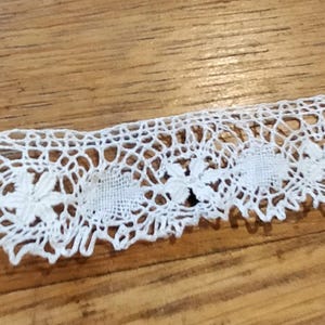 Peut inclure: Gros plan sur une bordure en dentelle blanche avec un motif floral. La dentelle présente un motif répétitif de motifs circulaires avec des détails floraux délicats et un bord festonné. La garniture est présentée sur une surface en bois.